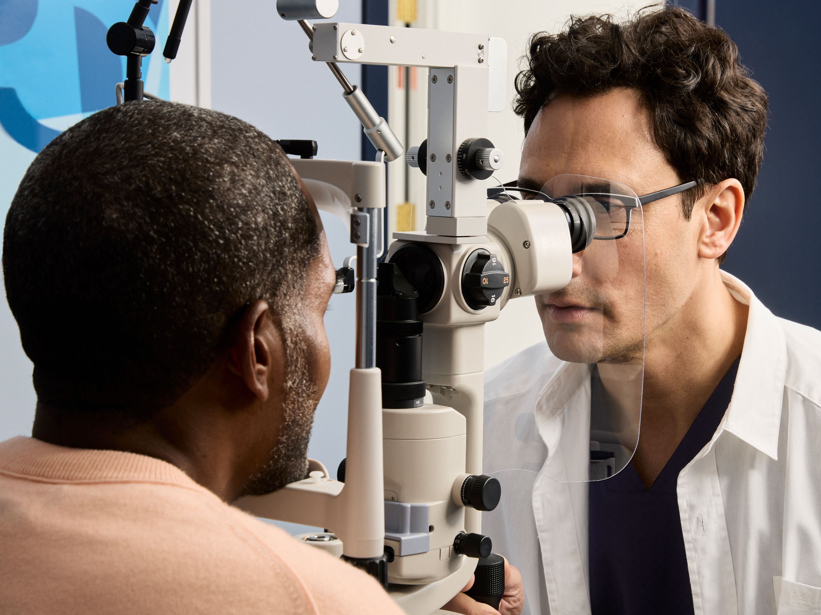 An eye exam in progress, with a patient looking into a slit lamp while an optometrist examines their eye closely.