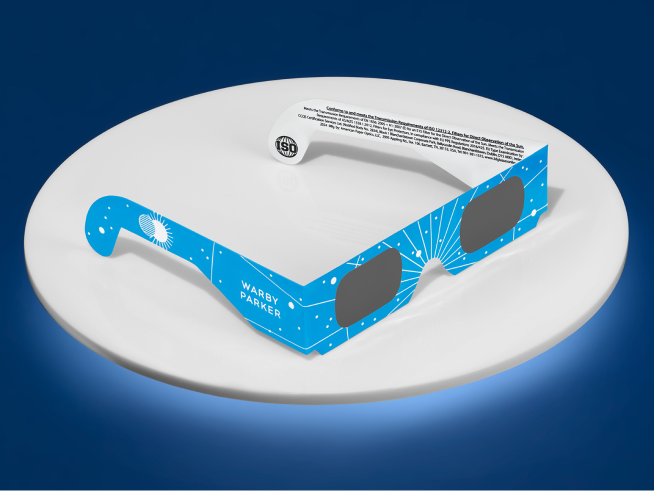 Blue eclipse glasses featuring a design with stars and rays, placed on a white circular dish against a dark blue background, with an instruction label visible on the upper side.
