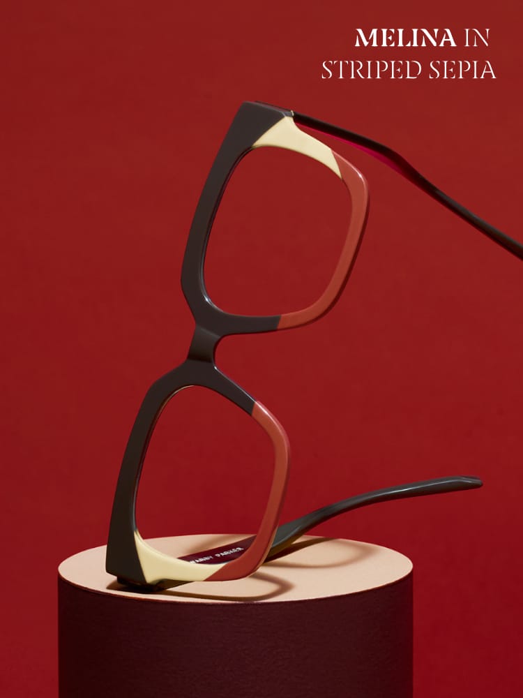 Unique eyeglasses with a geometric frame in a "striped sepia" color scheme, combining shades of black, red, and cream. The glasses are artistically balanced on a cylindrical pedestal against a deep red background. Text in the top right corner reads, "Melina in Striped Sepia," highlighting the eyewear's bold and artistic design.