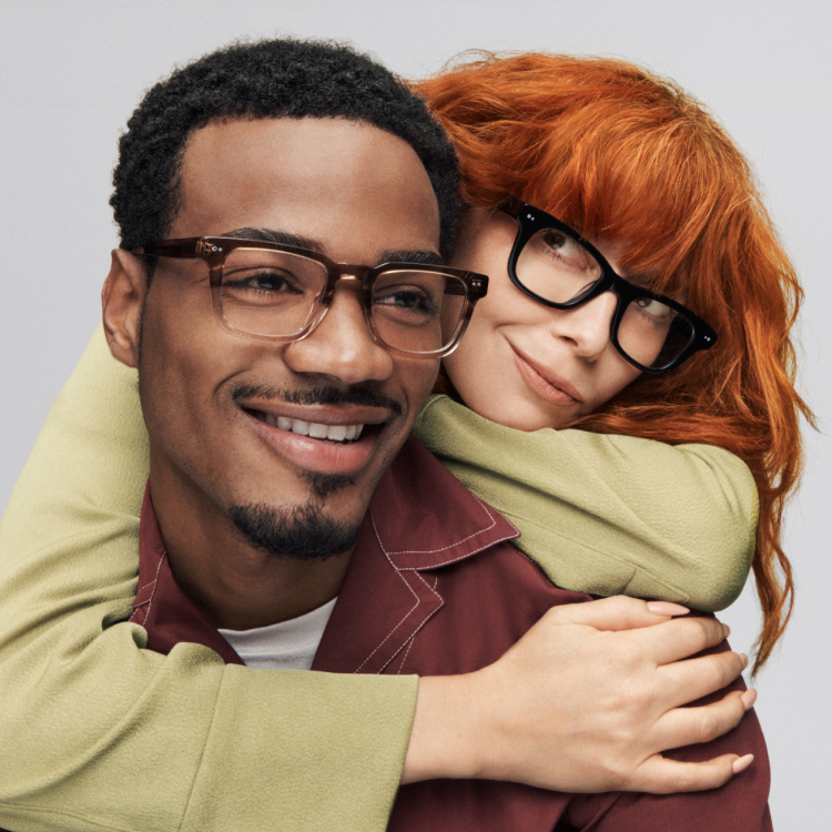 Natasha Lyonne embraces Tyshawn Jones from behind as they both sport stylish Warby Parker frames, sharing a lighthearted moment while smiling at the camera.
