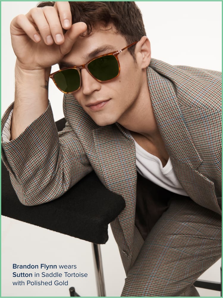 A person wearing sunglasses with a plaid suit, resting on a chair. Text: "Brandon Flynn wears Sutton in Saddle Tortoise with Polished Gold."
