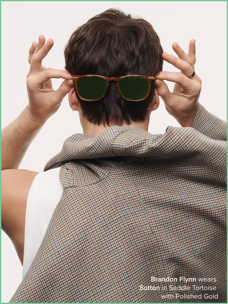 A person facing away, holding sunglasses behind their head, wearing a plaid jacket over their shoulder. Text: "Brandon Flynn wears Sutton in Saddle Tortoise with Polished Gold."