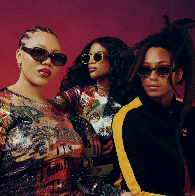 Three people wearing stylish sunglasses and colorful outfits, posing confidently against a dark red background.