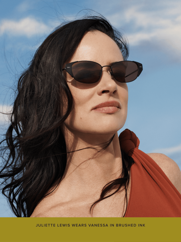Person with long black hair wearing dark sunglasses, looking confidently at the camera against a blue sky backdrop. Text below reads, "Juliette Lewis wears Vanessa in brushed ink."