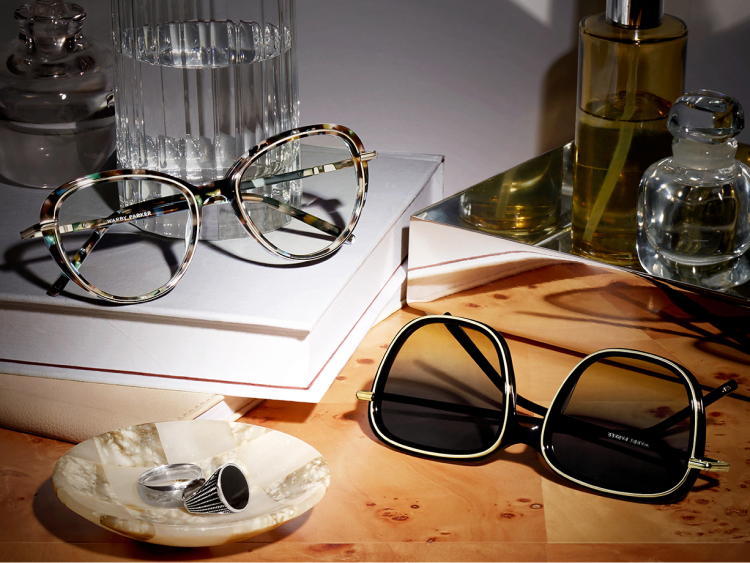Eyeglasses and sunglasses on a wooden surface, surrounded by books, a glass bottle, and a decorative dish with rings. Text: "Lorna in Aventurine Tortoise with Riesling and Safia in Jet Black with Polished Gold."