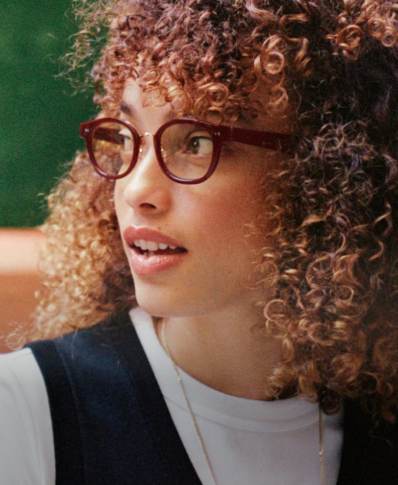 A young woman with curly, auburn hair and glasses looks to the side.