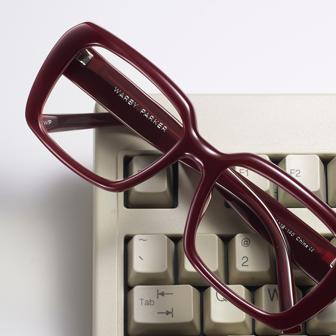Maroon Warby Parker glasses rest on a beige keyboard, with the brand name visible on the temple.