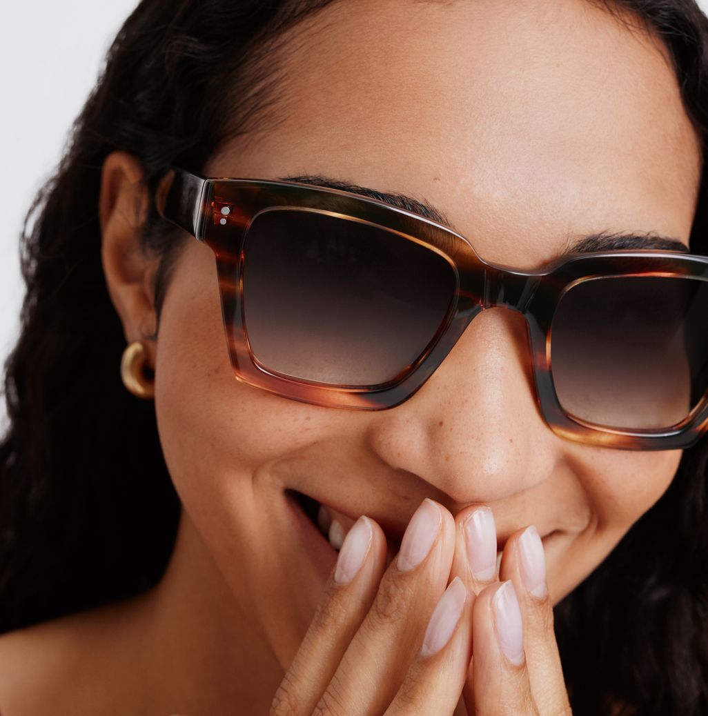 Close-up of a woman wearing oversized tortoiseshell sunglasses, laughing with hands covering her mouth.