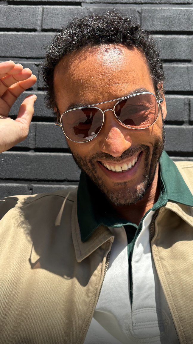 Man wearing sunglasses and a tan jacket with a green collar over a white polo shirt, smiling.