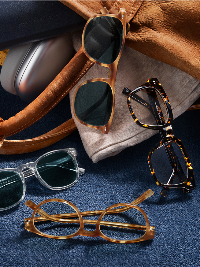 Four pairs of eyeglasses and sunglasses on fabric next to a brown leather bag and case.