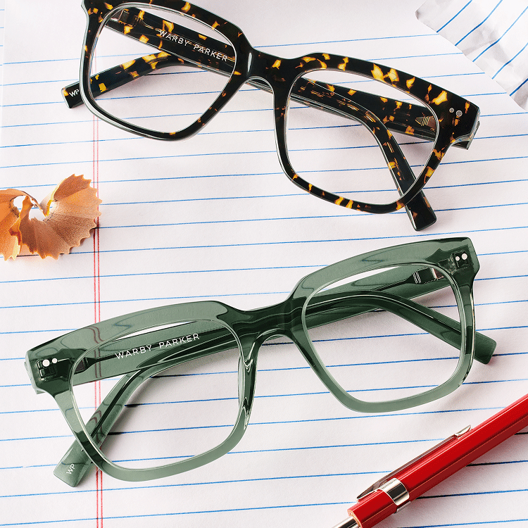 Two pairs of Warby Parker glasses on lined notebook paper with a red pen and pencil shavings.