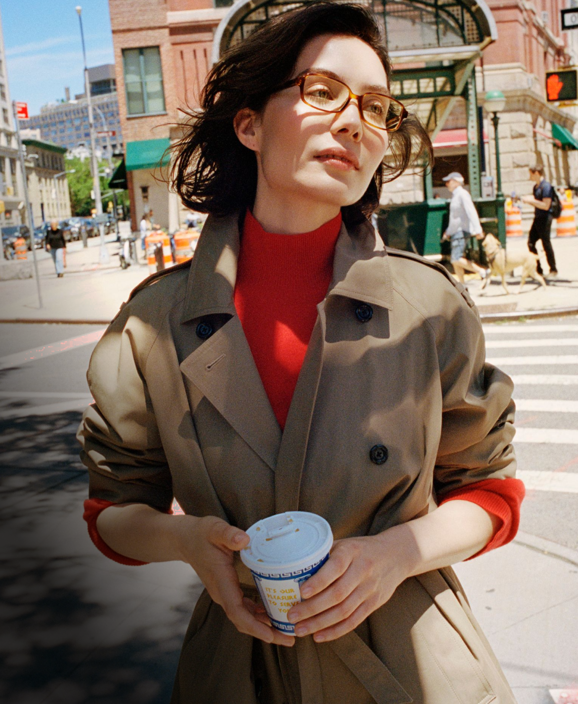 A woman in a trench coat and red turtleneck holds a coffee cup on a city street.