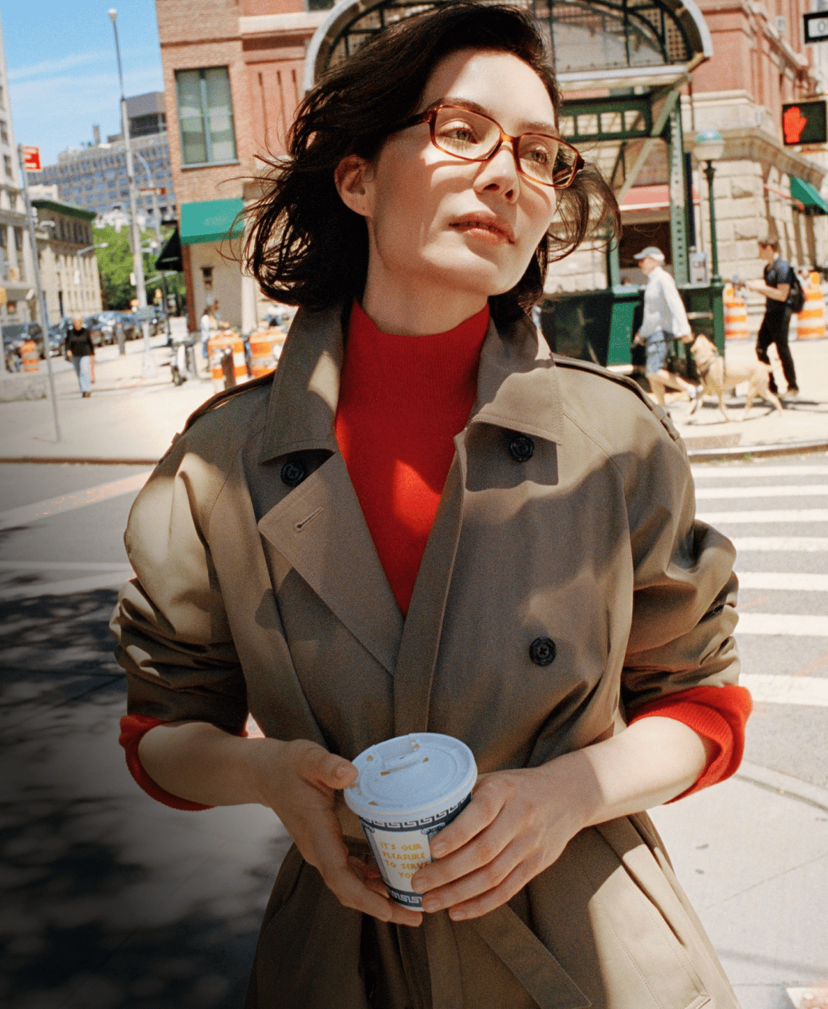 A woman in a trench coat and red turtleneck holds a coffee cup on a city street.