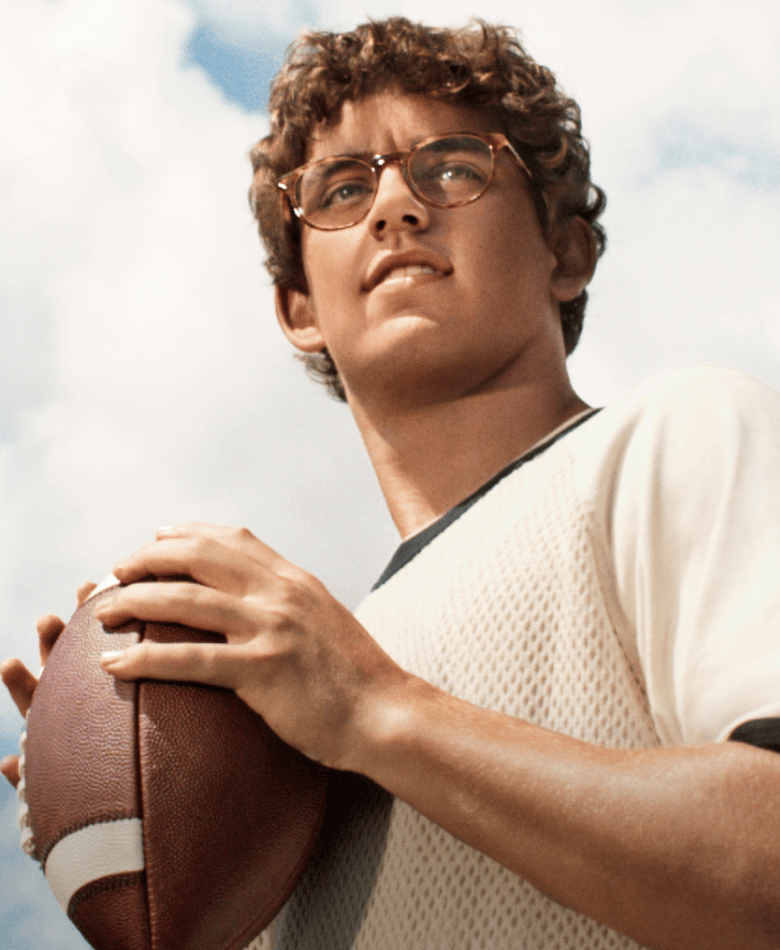A young man with curly brown hair and glasses holds a football in his hands, ready to throw.