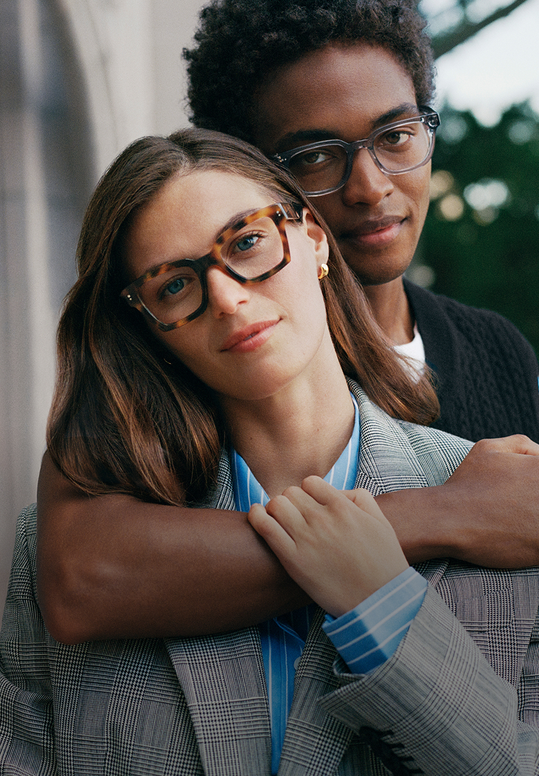 Couple outdoors wearing glasses, sitting closely with a green blurred background.