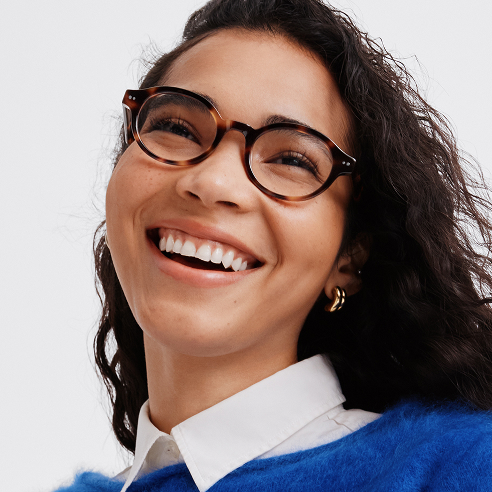 A smiling woman with dark curly hair wearing tortoise shell glasses and a blue sweater over a white shirt.