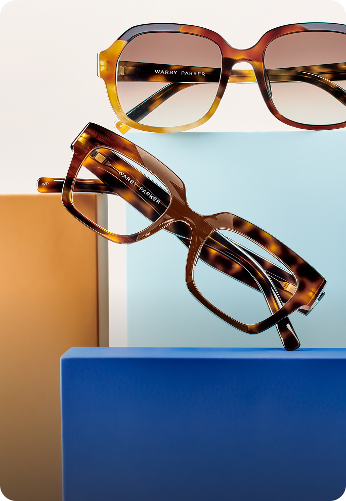 Two pairs of tortoiseshell Warby Parker glasses on blue and orange blocks with a light background.