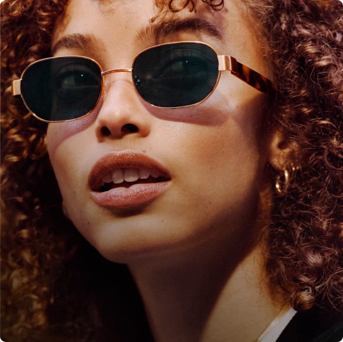 Close up of a woman with curly hair wearing sunglasses and a gold necklace.