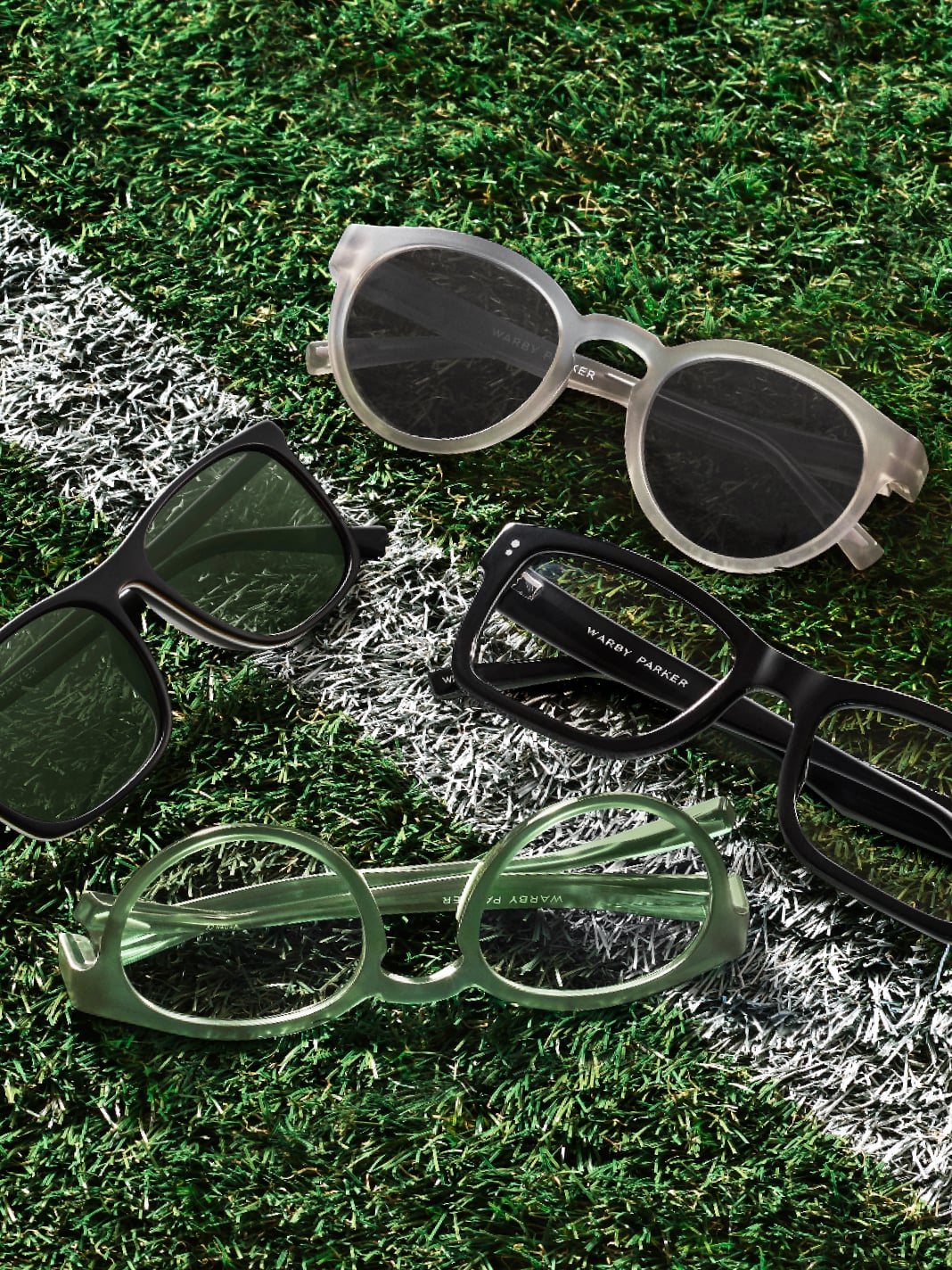 Four pairs of eyeglasses and sunglasses laid out on green artificial turf with a white line. The glasses include a pair of round translucent frames, black rectangular frames, black sunglasses, and translucent green oval frames.
