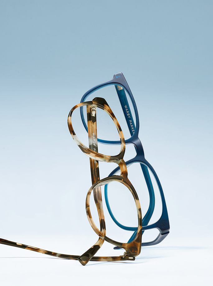 Two pairs of Warby Parker eyeglasses, one blue and one tortoiseshell, stacked against a light blue background.
