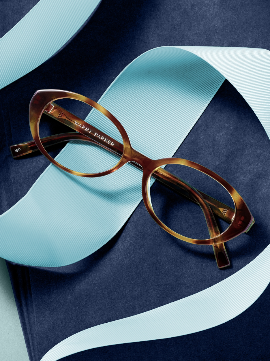 A pair of tortoiseshell cat-eye glasses rests on a navy background with a light blue ribbon.