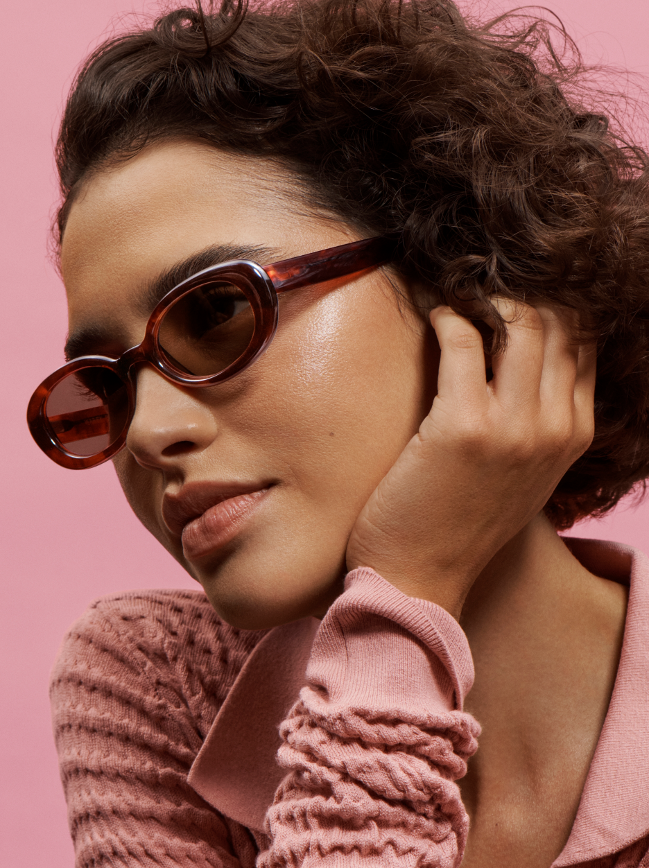 A woman with dark curly hair and brown sunglasses touches her face. She wears a pink sweater.