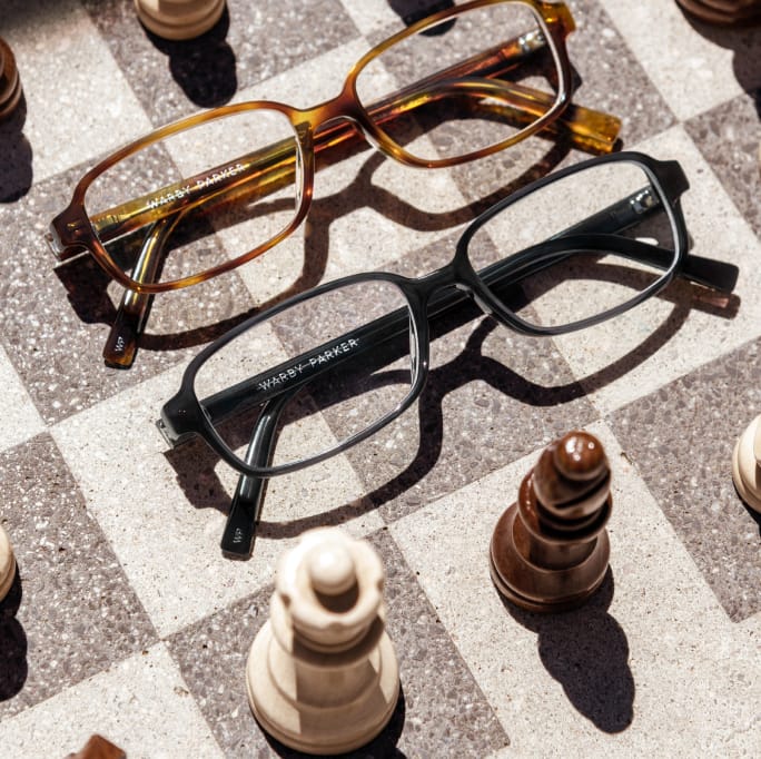 Two pairs of glasses, one tortoiseshell and one black, rest on a chessboard surrounded by chess pieces.