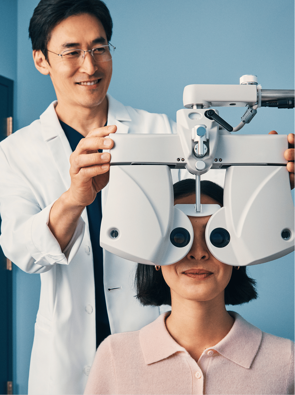 Person having their eyes examined with a phoropter by an optometrist against a blue background.