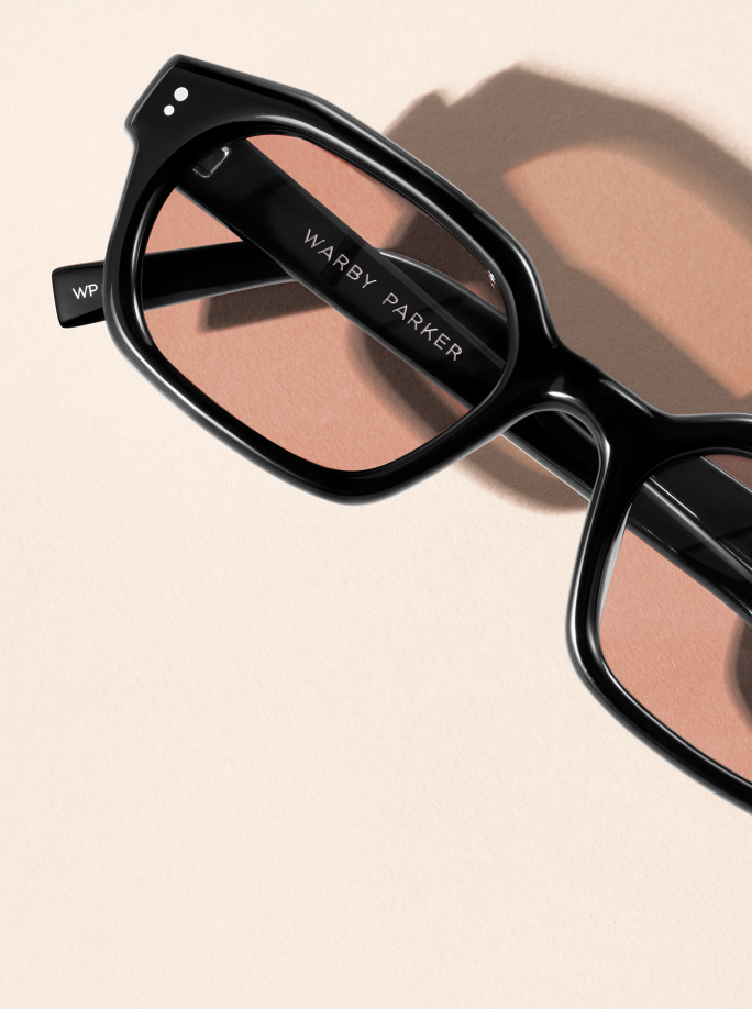 Black Warby Parker glasses with rose-tinted lenses casting a shadow on a light pink background.