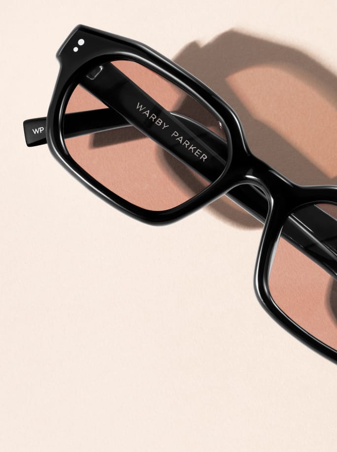 Black Warby Parker glasses with rose-tinted lenses casting a shadow on a light pink background.