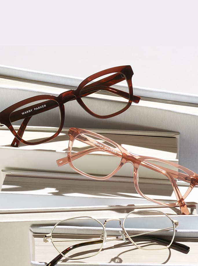 A woman in a beige jacket tries on a pair of eyeglasses, holding them up to her face while looking at a man in a black jacket standing in front of her, facing her.