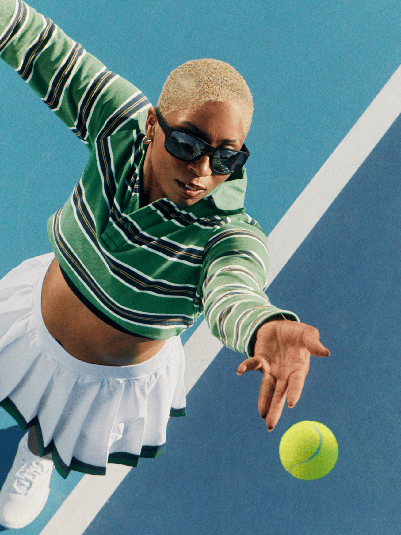 Woman in a green and white striped crop top and white tennis skirt reaching for a tennis ball.