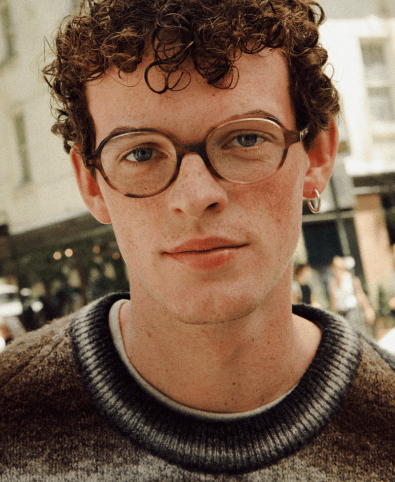 A young man with curly brown hair, wearing glasses and a sweater, looks into the camera.