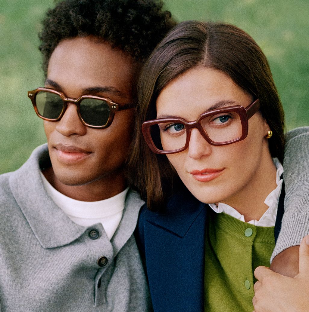 Man in sunglasses and woman in large brown glasses sitting closely outdoors.