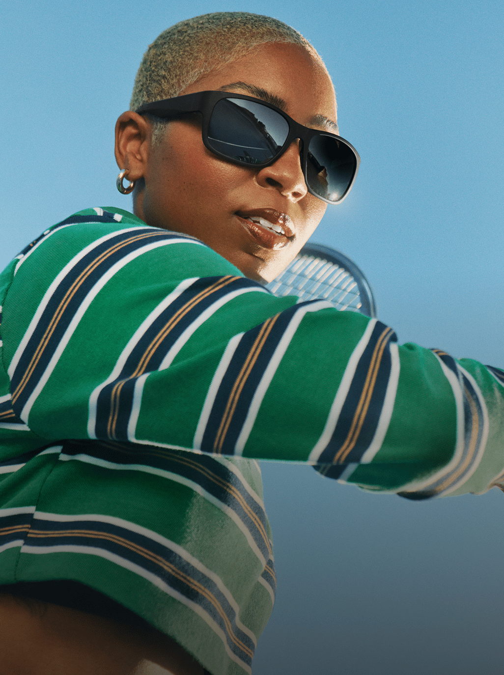 A woman with short, blonde hair and sunglasses wears a green striped polo shirt, holding a tennis racket.