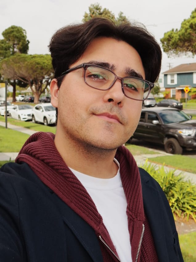 A young man with dark hair and glasses smiles for a selfie in front of a suburban street.