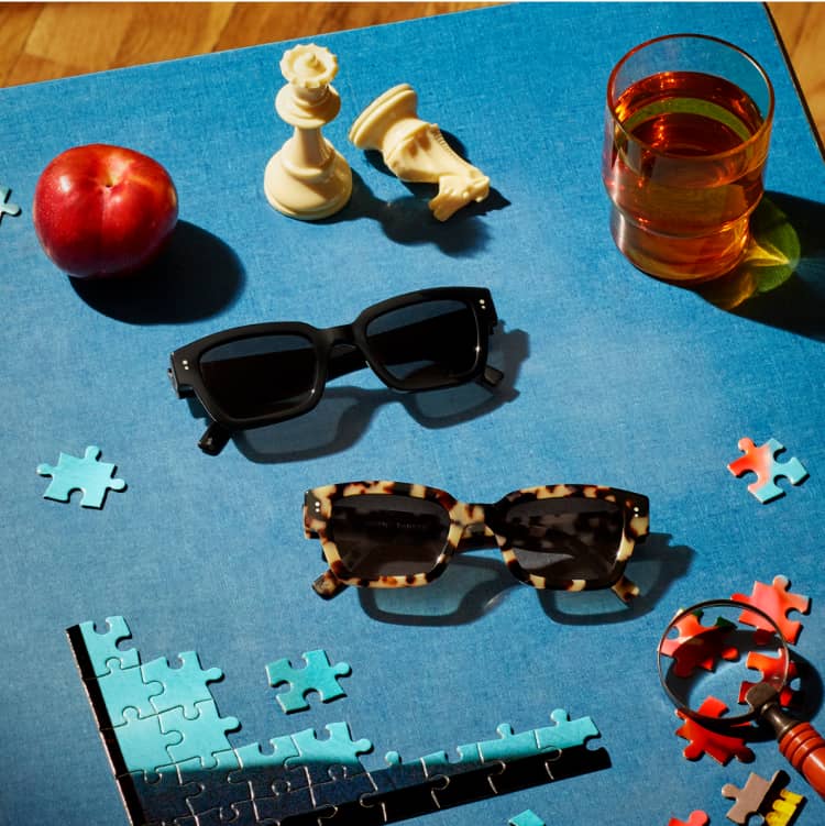 Acetate sunglasses placed on a blue mat surrounded by puzzles, chess pieces, apple, and a drink