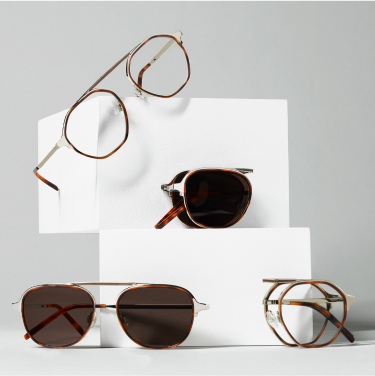 A stylish display featuring four pairs of glasses on stacked white blocks, including round and aviator sunglasses and sophisticated eyeglasses, all in various shades of brown and gold.
