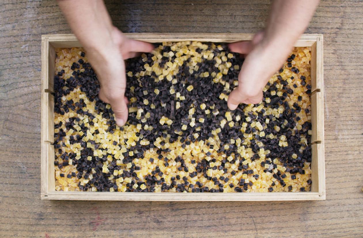 Mixed yellow and black cubed chips are placed into a wooden rectangle mold and mixed together by human hands. 