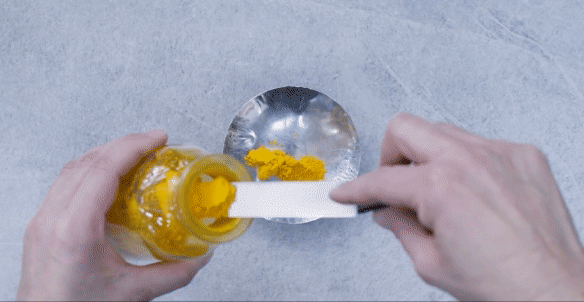 Looped video of Yellow powder pigment being scraped into a silver bowl, poured onto parchment paper onto of a marble white table, water being poured over yellow power, then folding the parchment paper in half mixing and spreading the yellow paste across the parchment paper. 