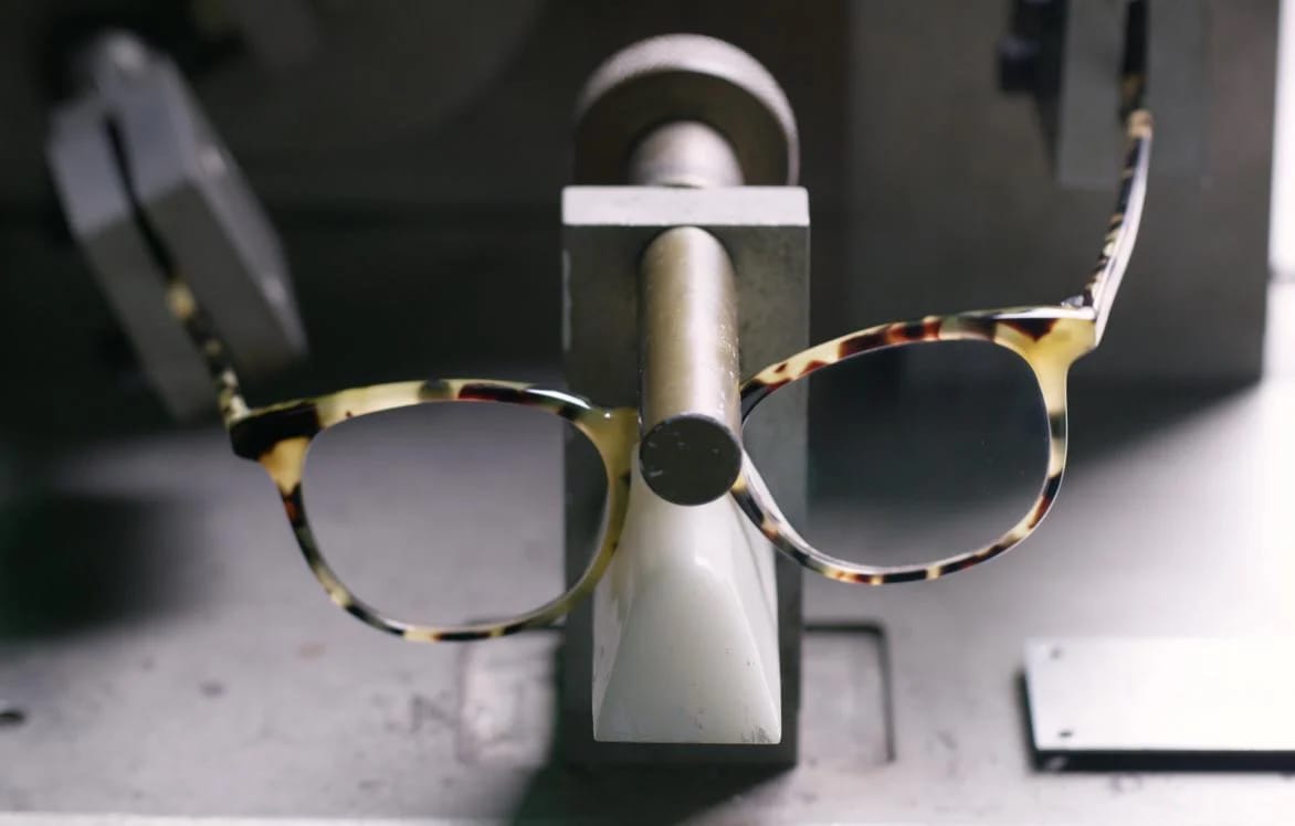Polished yellow and black tortoise acetate frame being held by their arms by a machine to bend and flex frame to test durability.