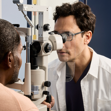 Male optometrist wearing rectangle eyeglasses and white medical coat performing eye exam on male patient. 