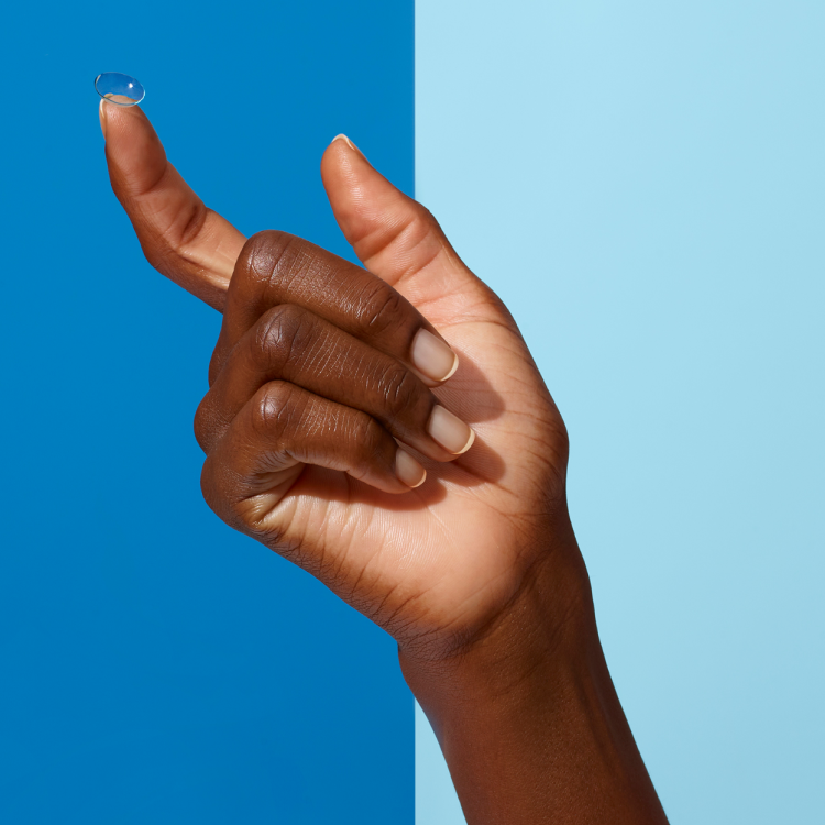 A hand holding a contact lens on the tip of the index finger against a blue background.