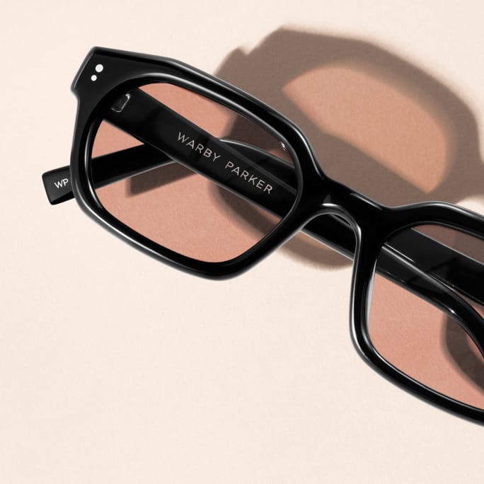 Black Warby Parker glasses with light brown lenses and shadow on a light pink background.