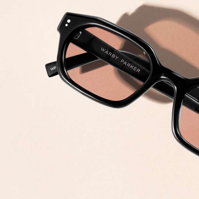 Black Warby Parker glasses with light pink lenses and a shadow on a pale pink background.