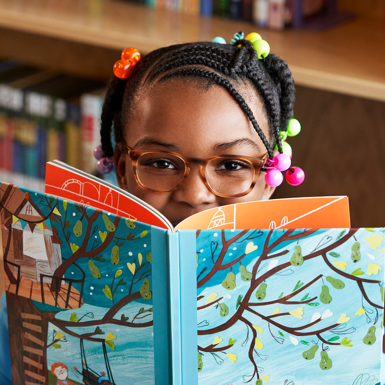 A young Black girl wearing glasses peeks over a colorful children's book in a library.