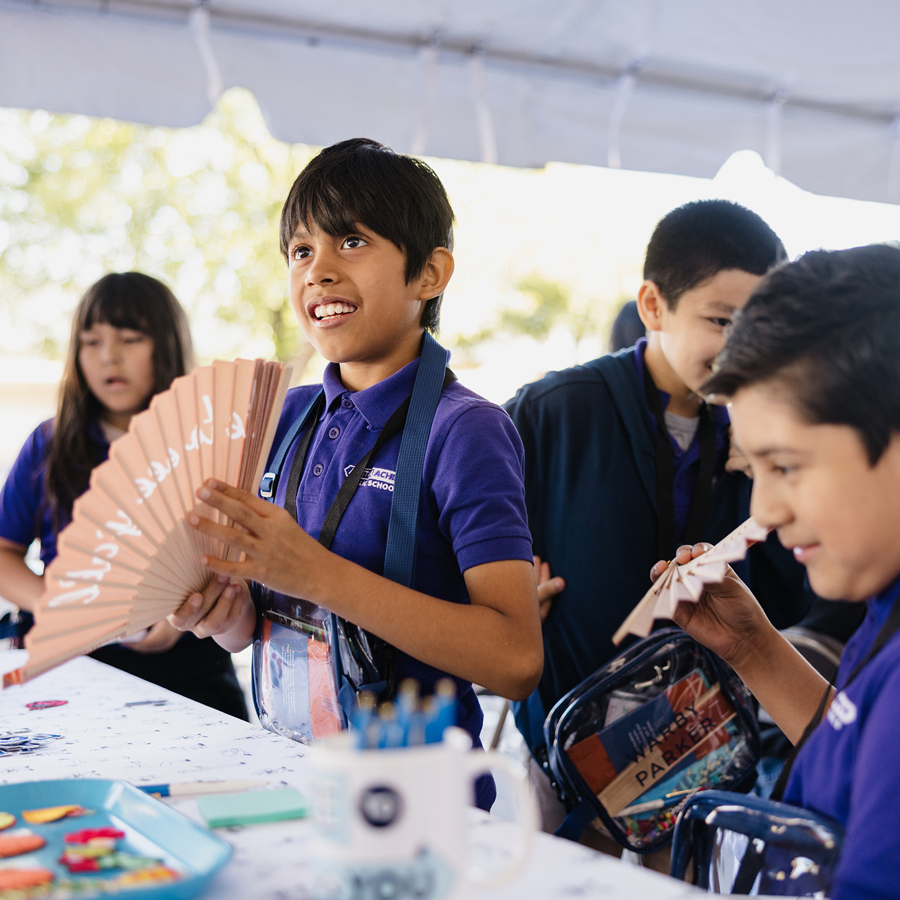 A boy in a purple shirt holds a fan. He smiles and looks to his left.