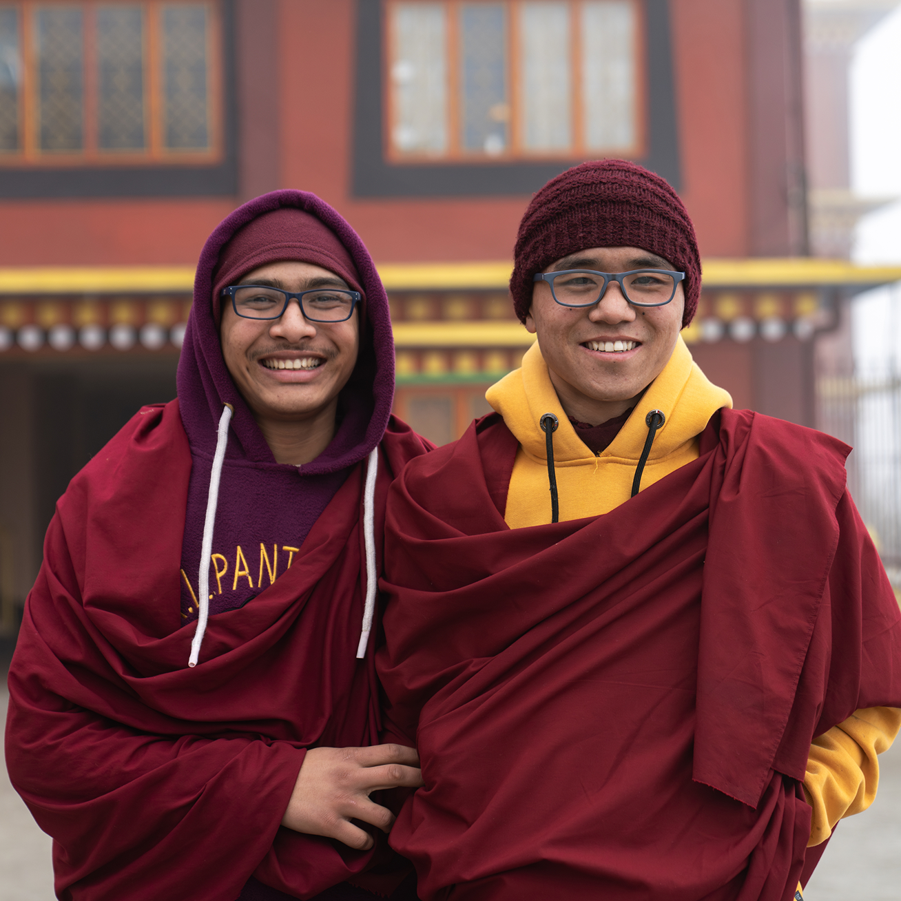 Two young Buddhist monks smile at the camera. They are wearing traditional maroon robes over hoodies.