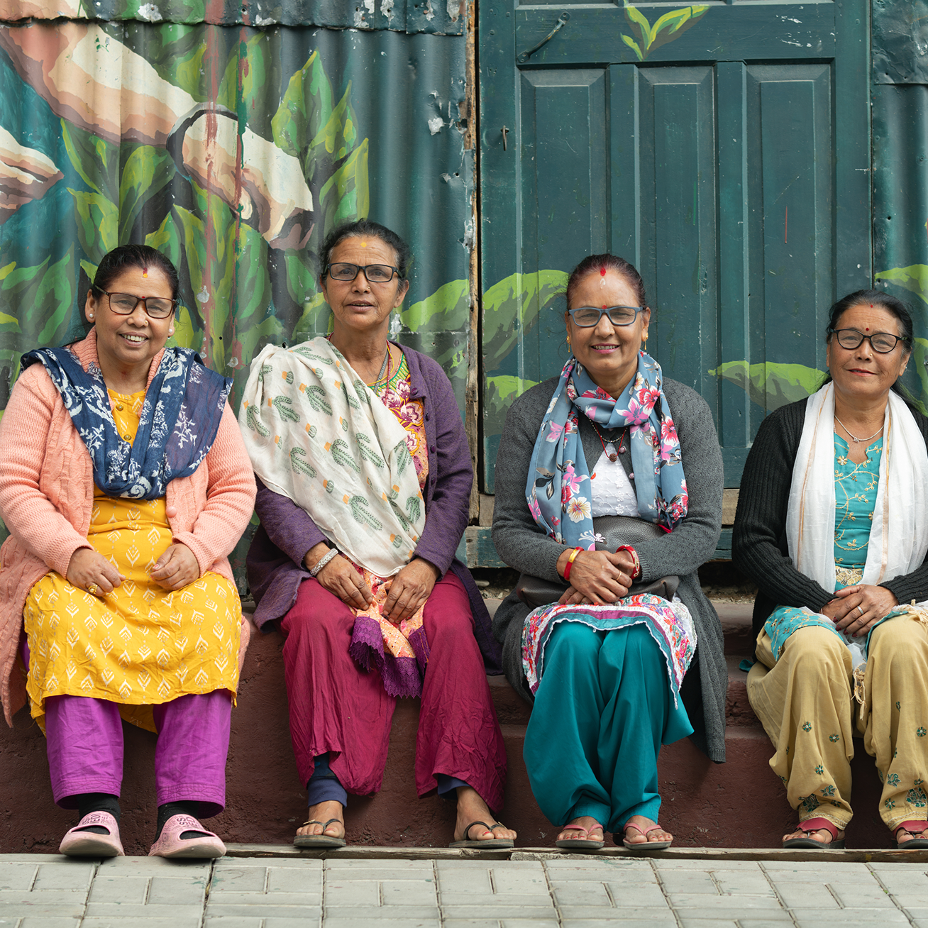 Two women sit on steps, smiling and chatting; one wears glasses and a pink scarf, the other also has glasses and is in a green apron with a phone in hand.