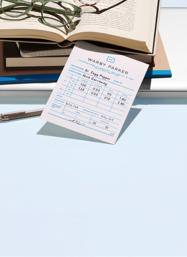 Eyeglass prescription slip on a desk, with open books in the background.